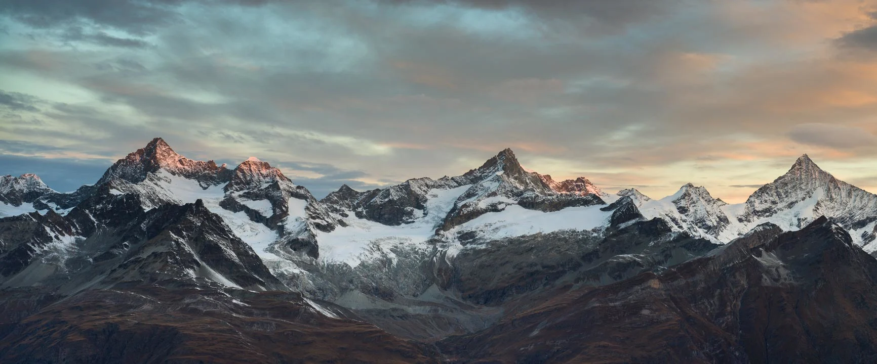 A panoramic shot of snow-capped mountain peaks under a cloudy sky at sunset.