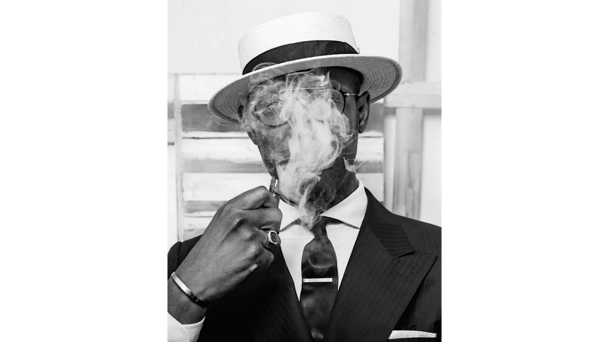 Photo en noir et blanc d'un homme élégant portant un chapeau et fumant. De la fumée s'échappe devant son visage.