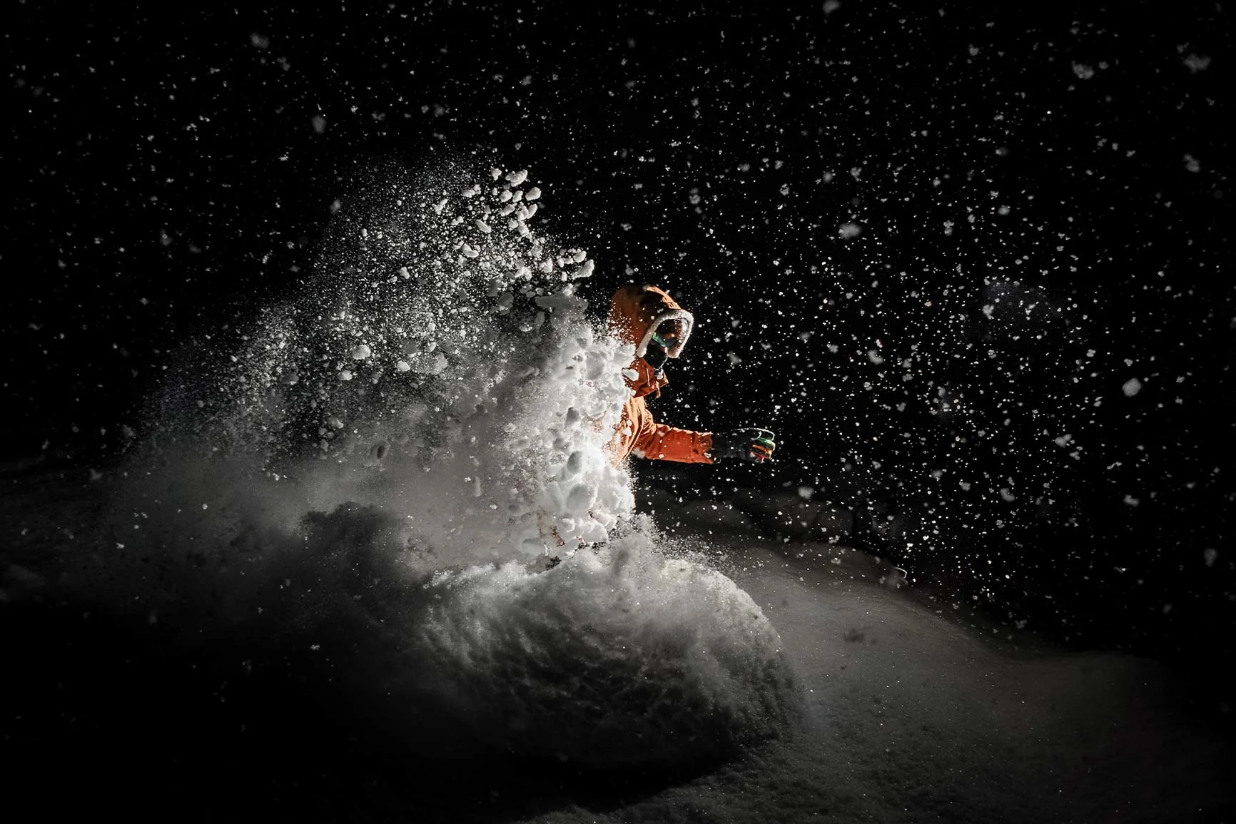 A skier wearing an orange jacket and goggles is skiing through deep snow against a black background.