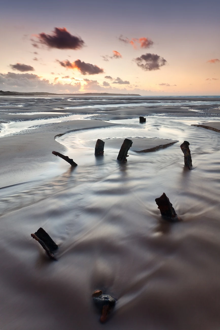 A deserted beach in front of a sunset at low tide, metal parts sticking out of the sand.