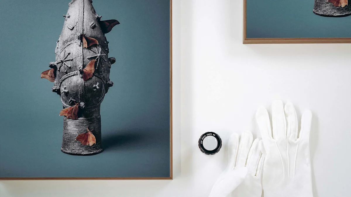 Die Draufsicht zeigt zwei gerahmte Kunstwerke auf einer weißen Arbeitsfläche, ergänzt durch professionelle Utensilien zur Kunsthandhabung. Auf der linken Seite ist ein großer Ausschnitt des vertikalen Motivs mit dem historischen Bronzeobjekt und den Faltern zu sehen, während oben rechts ein weiteres gerahmtes Bild angeschnitten ist. Im rechten unteren Bereich liegen ein Paar weiße Baumwollhandschuhe und eine kleine, schwarze Messlupe bereit. Die saubere, strukturierte Anordnung vermittelt die präzise und sorgsame Qualitätskontrolle oder Vorbereitung einer Ausstellung.