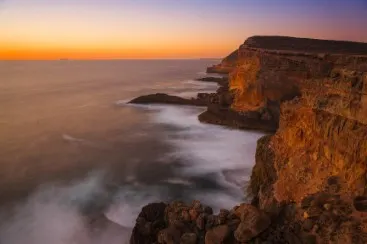 Cliff coast with rocks by the sea at sunset, calm water and orange evening light.