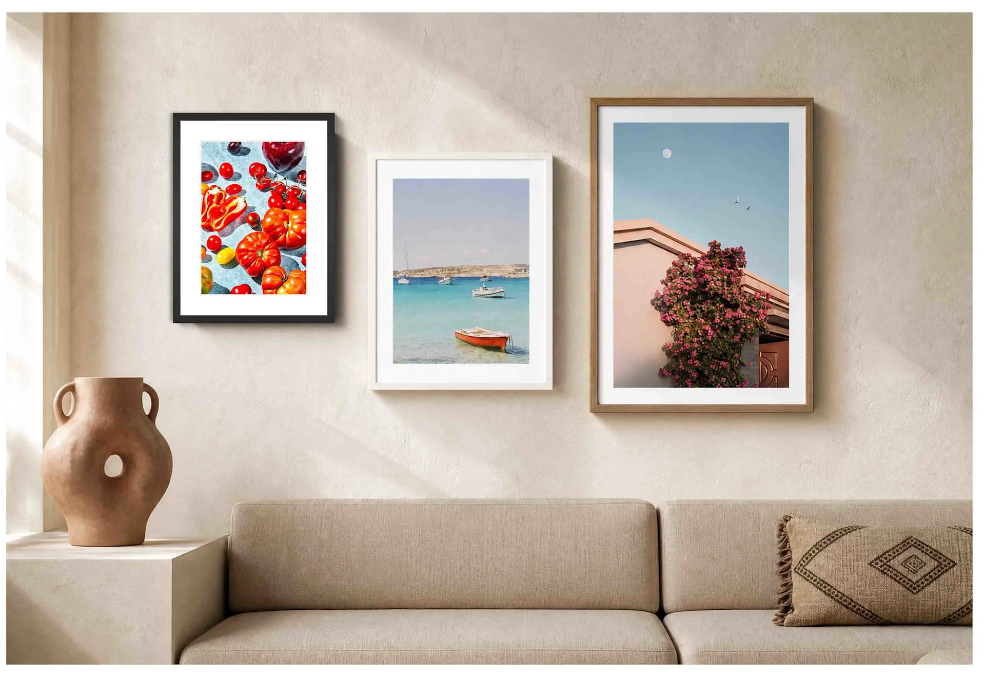 Un salón minimalista presenta un cómodo sofá de tela beige adornado con un cojín bordado con motivos marrones. En la pared beige del fondo, se alinean tres impresiones artísticas verticales enmarcadas. De izquierda a derecha, muestran un surtido de tomates llenos de color en un marco negro, una barca roja en aguas turquesas en un marco blanco y una fachada rosa bajo la luna llena en un marco de madera. Un gran jarrón abstracto de cerámica marrón con un hueco central descansa sobre el saliente de la izquierda bajo una luz suave.