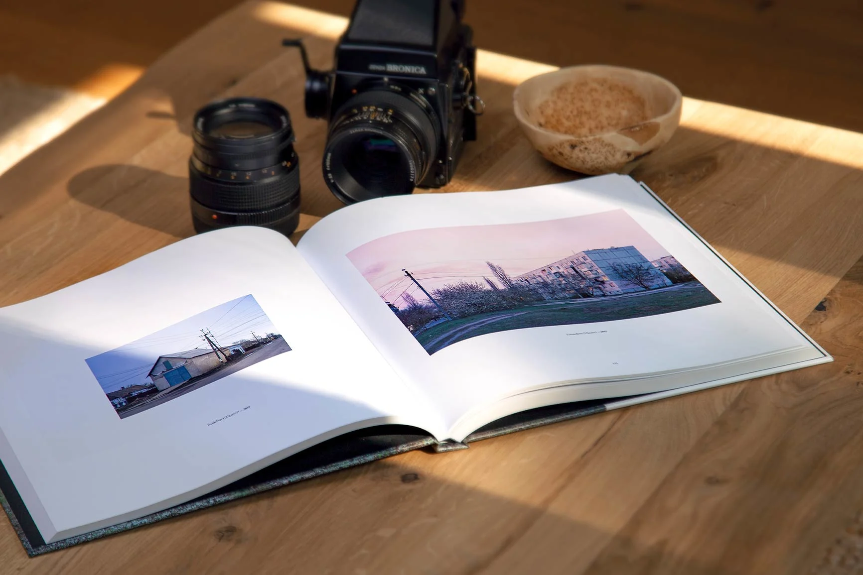 Aufgeschlagener Bildband mit Landschaftsfotos, daneben eine Mittelformatkamera mit Objektiv und eine Schale auf einem Tisch.