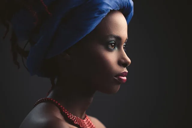 A woman in profile is wearing a blue headscarf and a red necklace. She has blue eye shadow and red lips, the background is dark.