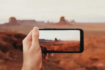 Mano sosteniendo un smartphone, mostrando un paisaje desértico con rocas y suelo rojo, fondo desenfocado.