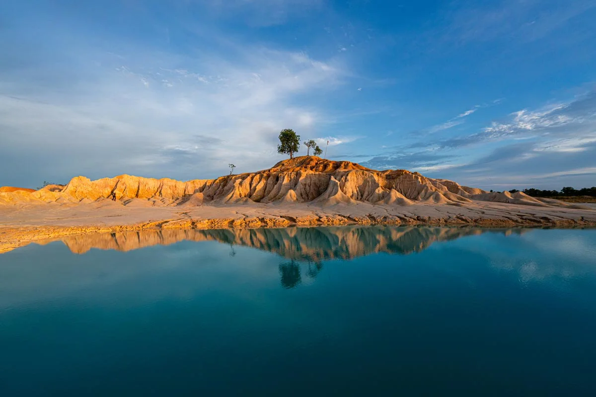 A sandy hill is reflected in a calm, blue body of water. There are a few trees on the hill, the sky is blue with light cloud formations.