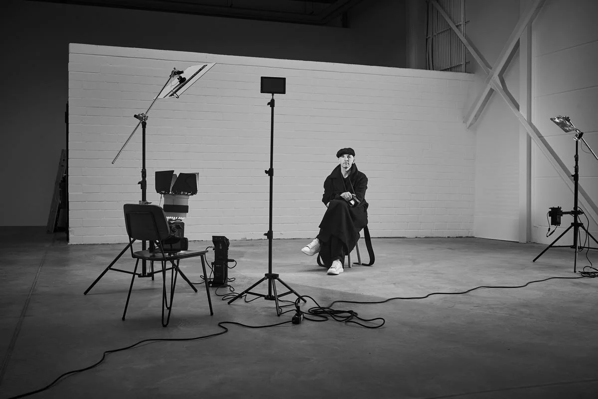 Black and white photograph of a studio with a person sitting on a chair. The person is wearing a coat, a cap and white shoes. Around them are several lighting devices, tripods and a chair with equipment. The back wall of the studio is made of white bricks and cables are scattered on the floor.