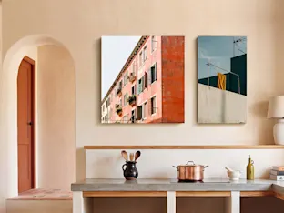 Interior de cocina minimalista con dos impresiones artísticas verticales montadas en una pared beige de tono cálido sobre una encimera de hormigón. La impresión cuadrada más grande a la izquierda muestra la fachada de un edificio de terracota de varios pisos con ventanas y jardineras, mientras que la impresión rectangular más pequeña a la derecha presenta una toalla amarilla colgada de una cuerda ante una estructura verde azulado. Debajo de las obras, una olla de cobre sobre una placa de cocción, un mortero con mazo y utensilios de madera en un tarro de cerámica decoran la encimera. Un arco a la izquierda se abre a una habitación contigua con suelo de baldosas de terracota.