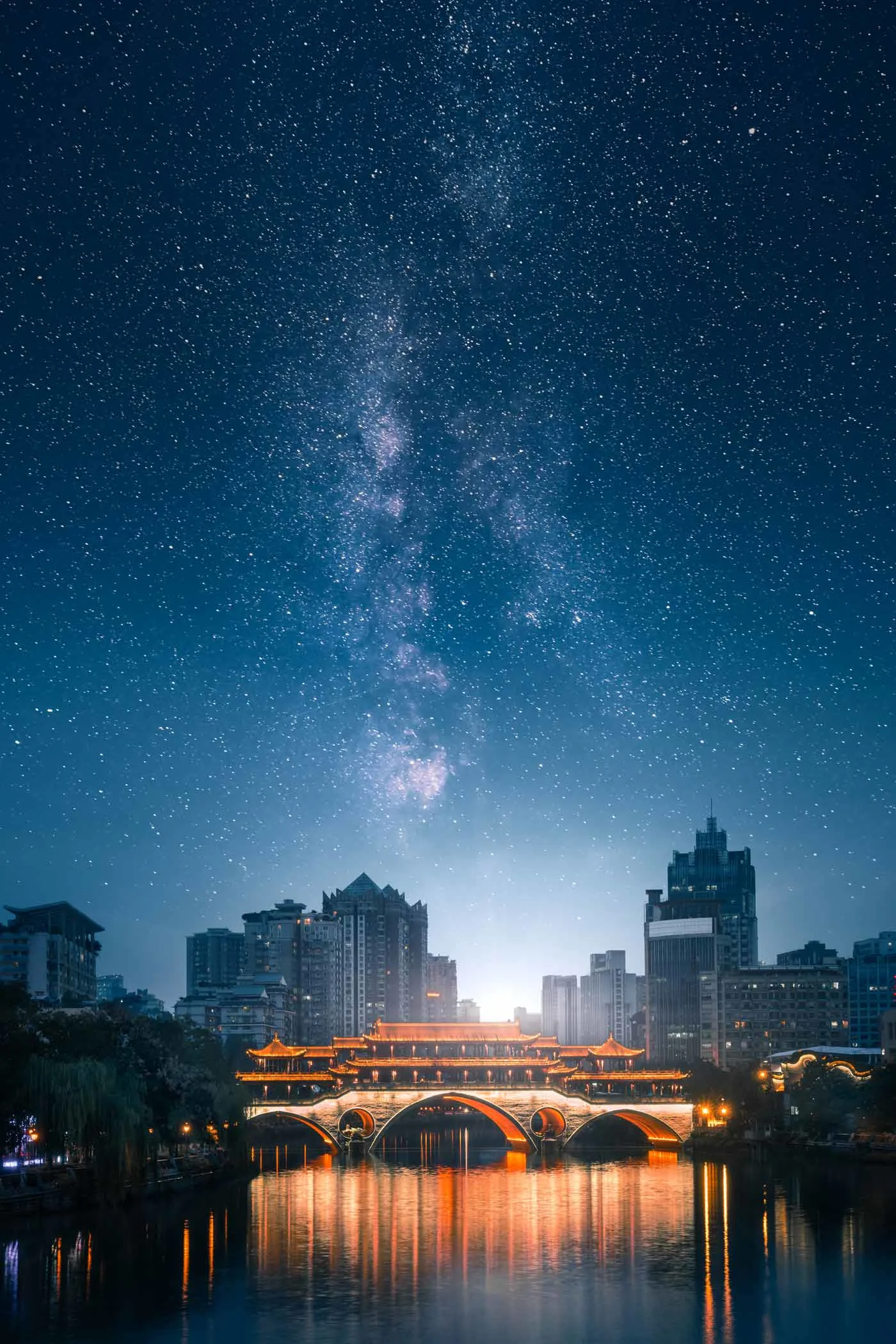 Night shot of a city with an illuminated bridge over a river in the foreground. The starry sky with the Milky Way is visible above the skyscrapers.