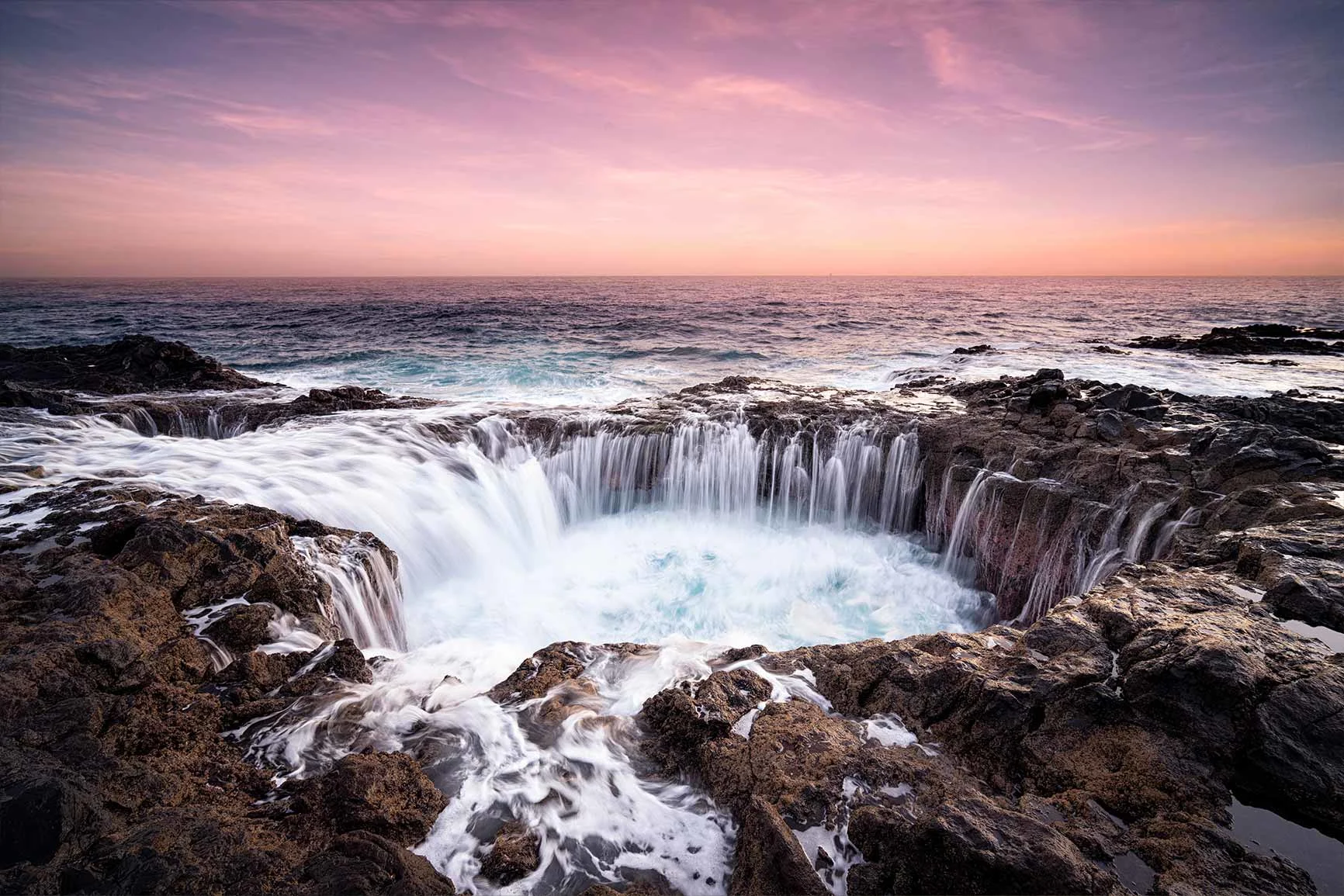 Water flows over a rock formation by the sea into a circular hole, sunset on the horizon.
