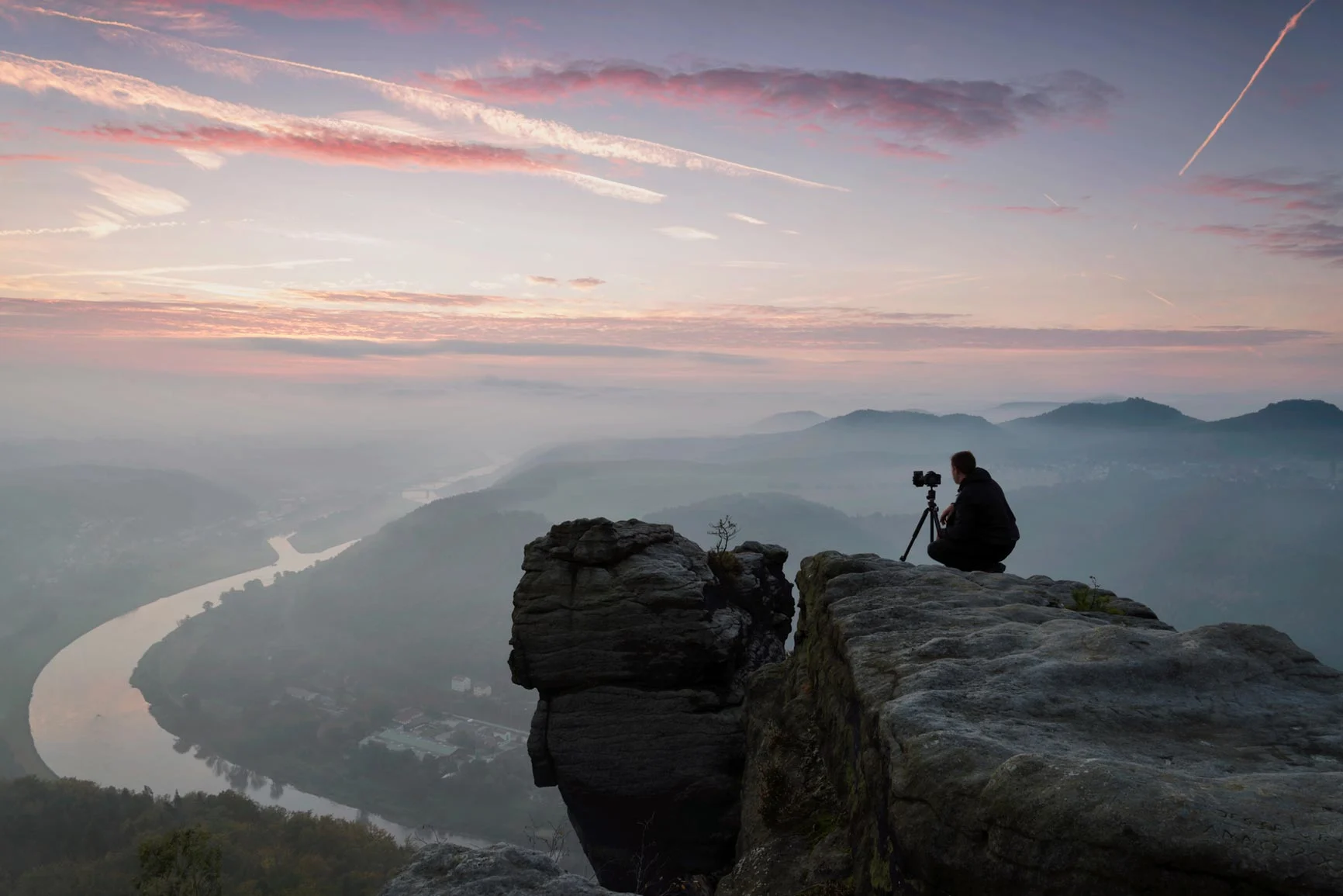A man is sitting on a rock with a camera mounted on a tripod, looking out at a misty river in the valley at sunrise.