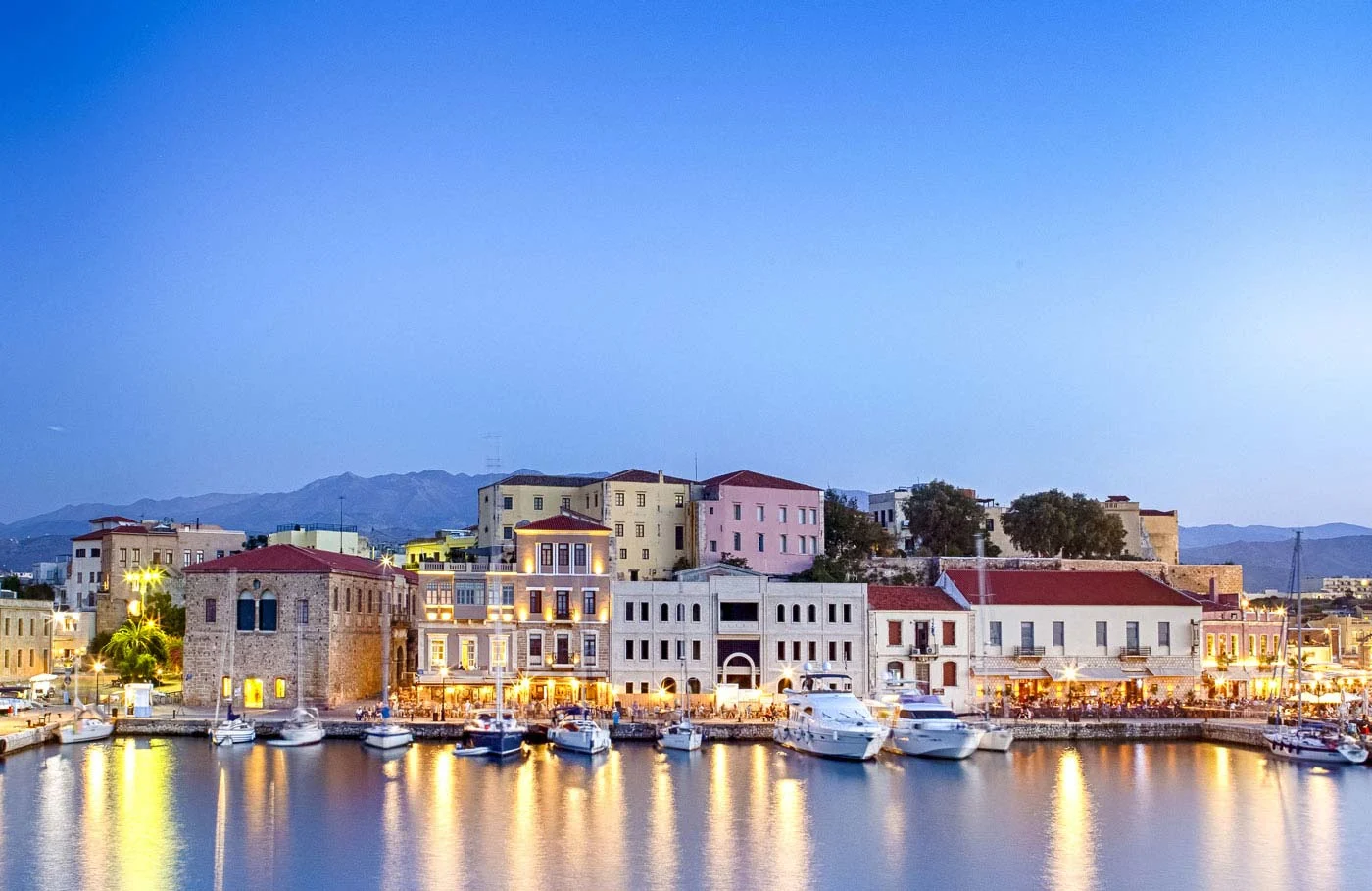 Harbor with illuminated buildings and boats against a mountain backdrop at dusk.