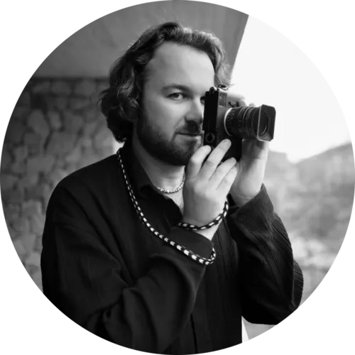 Ein kreisförmig zugeschnittenes Schwarz-Weiß-Porträt zeigt den Fotografen Alan Schaller. Er blickt direkt in die Kamera, während er eine Leica-Kamera vor sein Gesicht hält, so als würde er gerade ein Foto machen. Er trägt ein dunkles, strukturiertes Hemd und eine markante, gemusterte Kameraschlaufe ist um sein Handgelenk gewickelt. Der Hintergrund zeigt eine unscharfe Außenansicht mit einer Steinwand.