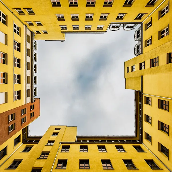 A dramatic worm's-eye view looking straight up into the square courtyard of a modern building. The four inner walls are painted bright yellow and feature numerous evenly spaced windows. At the top, the roof edges form a precise frame around a patch of gray, cloudy sky. A single section of wall in the lower area features red bricks.