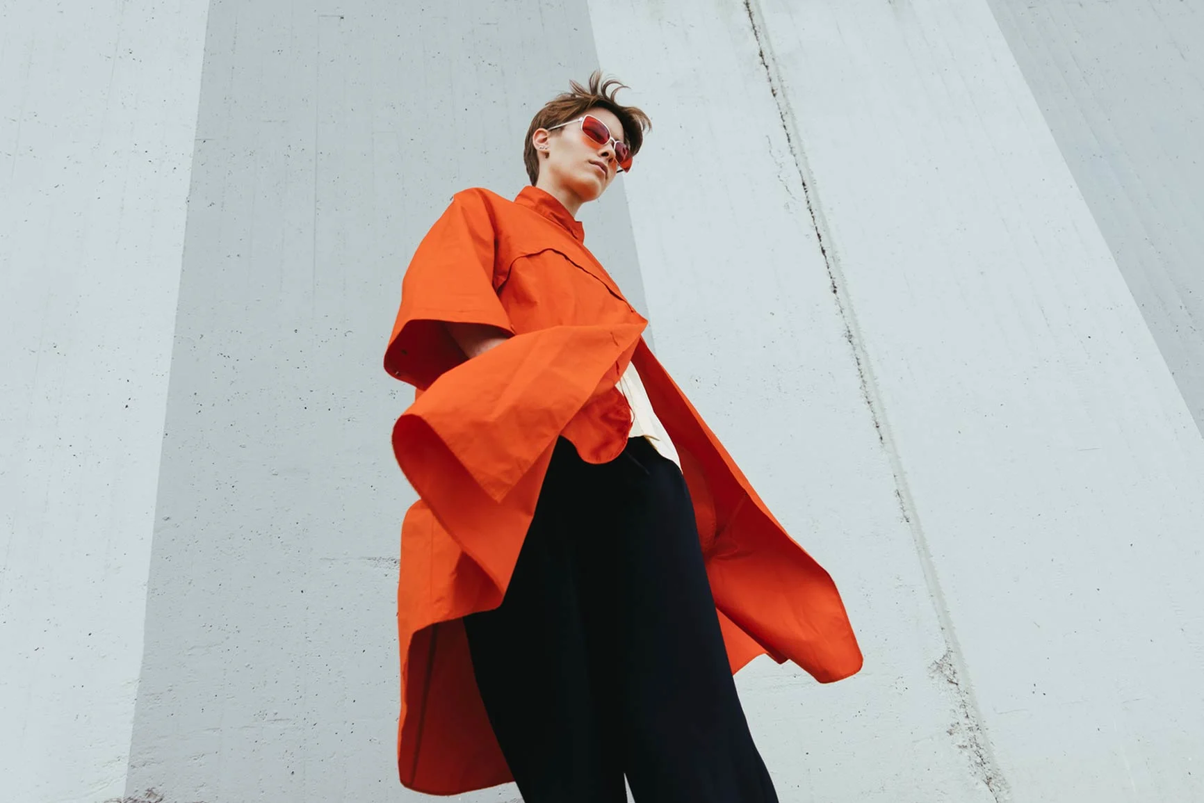 A woman with short hair is wearing a loose-fitting orange coat and black pants in front of a light gray wall.