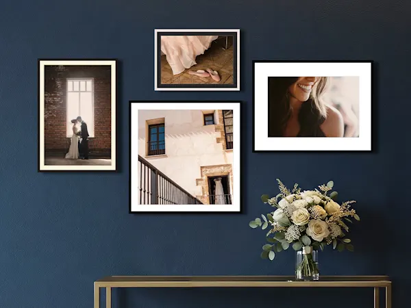 Four framed wedding photos on a dark blue wall above a gold console table with a white rose bouquet.