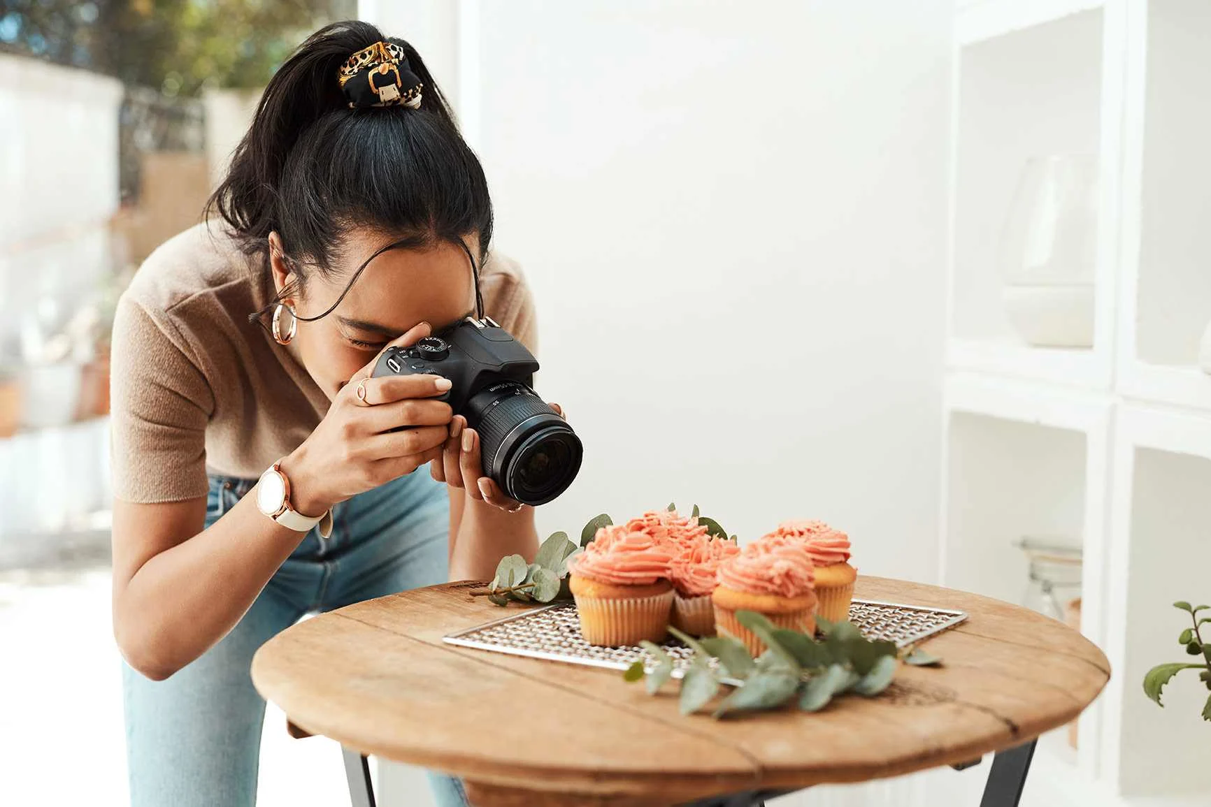 A woman is leaning over a round wooden table and taking pictures of three cupcakes decorated with pink frosting.