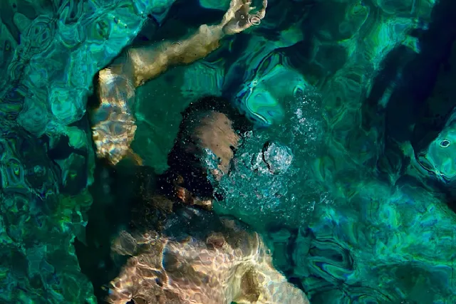 Man swimming underwater with arms outstretched, water surface showing light reflections and shades of green and blue.