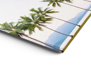 Une impression sur toile avec bords repliés, représentant de grands palmiers sur fond de ciel pâle et d’eau bleue de l’océan, présentée en biais sur une surface blanche.