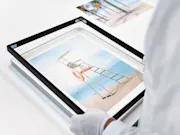 Une personne portant des gants change le cadre d'une image magnétique encadrée. L'image montre une scène de plage avec une chaise haute pour un maître-nageur.
