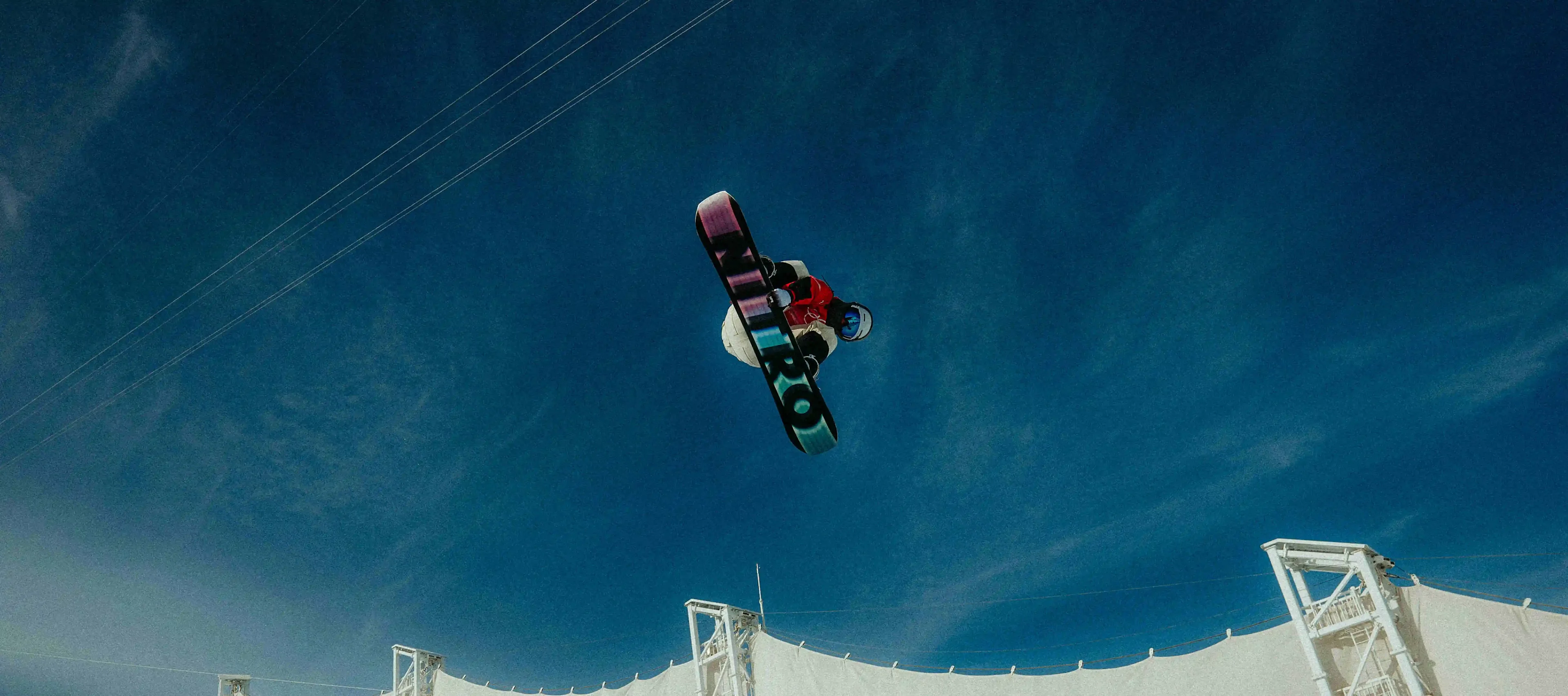 A snowboarder performs a high jump against a deep blue sky. The underside of the snowboard is visible, featuring the prominent "NITRO" branding. Diagonal power lines or cable car wires run through the background, emphasizing the dynamic motion.
