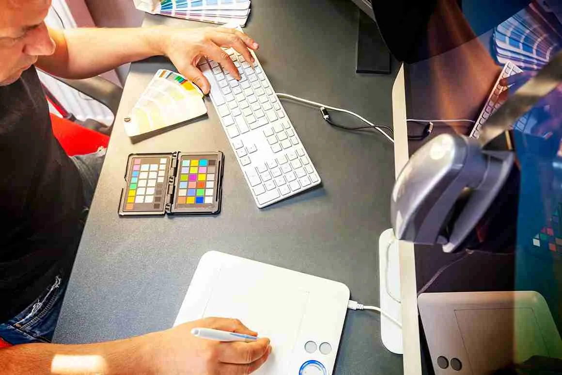 Mann, der an einem Schreibtisch an einem Grafiktablett arbeitet, mit einer Tastatur, einer Maus und Farbmuster vor sich.