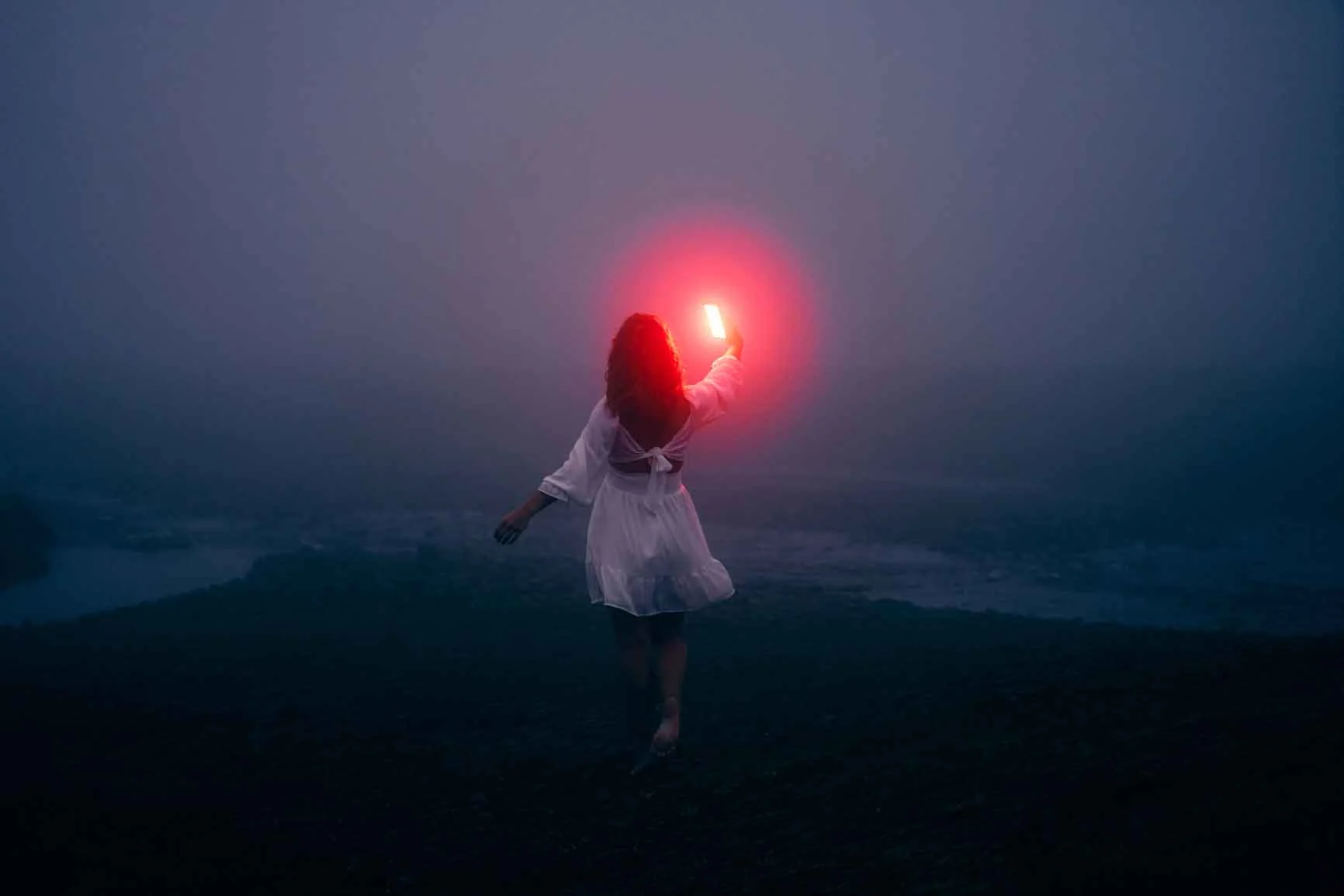 A person in a white dress stands in the fog and holds a bright red light in her outstretched hand. The background is dark and foggy.