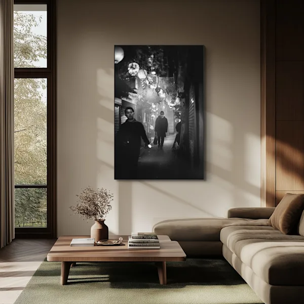 Ein helles, modernes Wohnzimmer zeigt ein großformatiges, vertikales Schwarz-Weiß-Foto an der Wand. Das Motiv von Phil Penman zeigt eine nächtliche Straßenszene in einer schmalen Gasse mit Passanten und leuchtenden Lampions. Unter dem Bild steht ein niedriger Holztisch mit Büchern und einer Vase, daneben ein großes, beige-braunes Ecksofa. Sonnenlicht fällt durch ein großes Fenster und wirft Schattenstreifen an die Wand.