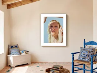 Escena d'interior elegante y luminosa con una gran impresión de retrato vertical centrada en una pared beige de tono cálido. La obra de arte, presentada dentro de un marco magnético de aluminio blanco de alta gama con un amplio passe-partout blanco, muestra un primer plano del retrato de una persona con cabello rubio bajo la luz directa del sol que proyecta sombras alargadas. Debajo de la obra, un suelo de baldosas de terracota rústico está decorado a la izquierda con un baúl blanco con cojines azules y a la derecha con una silla de madera azul brillante con cojines geométricos a juego. Una mesa de centro de madera baja con una pila de libros antiguos se encuentra en el centro del primer plano.
