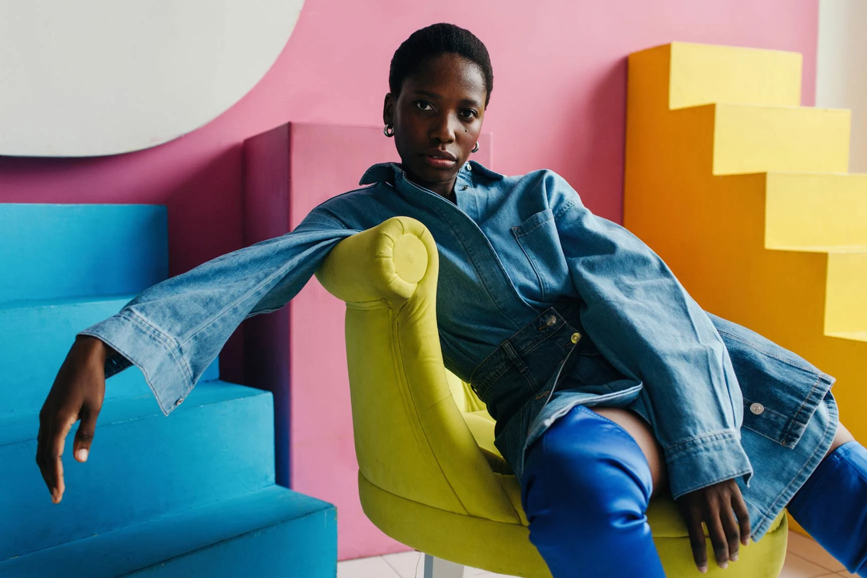 A woman wearing a denim jacket and blue pants is leaning back comfortably in a yellow armchair in front of colorful geometric shapes.