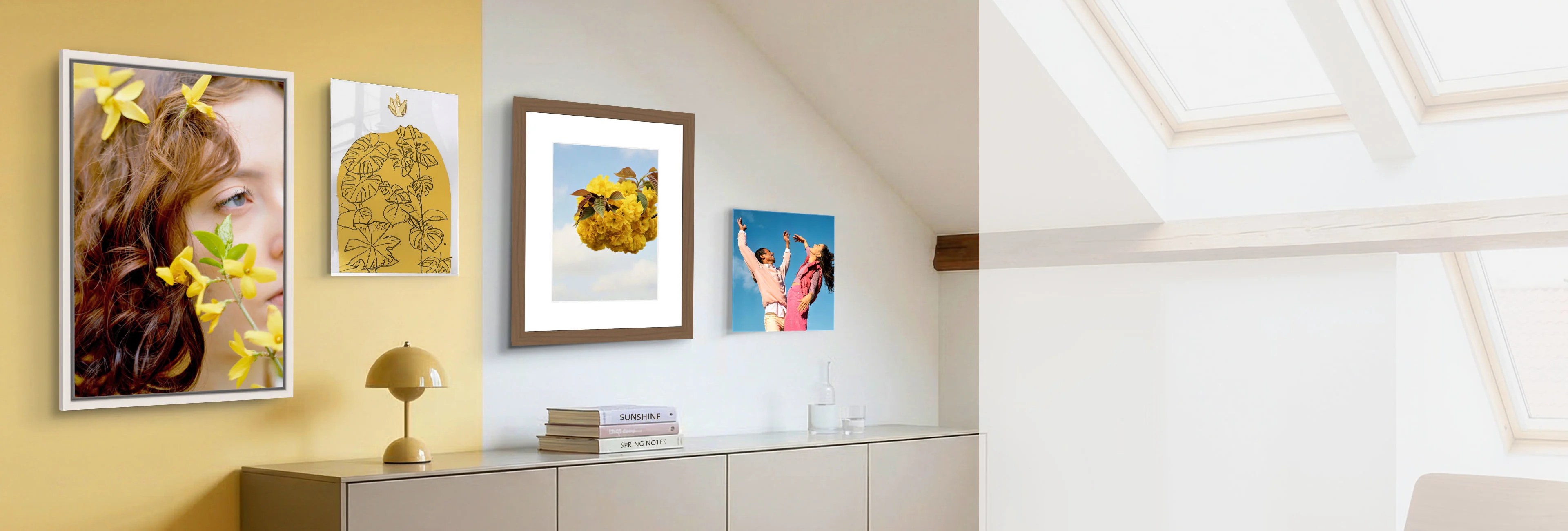 Una vista di una stanza luminosa e accogliente sotto un soffitto inclinato con ampi lucernari. Sulla parete sopra una lunga credenza grigia sono appese diverse presentazioni d'immagine: un ritratto in una cornice bianca a cassetta, una stampa su metallo lucida, un motivo floreale incorniciato con passe-partout e un pannello fotografico a montaggio diretto. Una lampada di design gialla e una pila di libri sulla credenza completano perfettamente l'ambiente moderno.