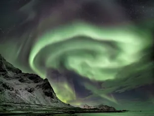 Vivid green aurora borealis swirling in a spiral over snow-dusted mountains and a calm coastal shoreline at night.