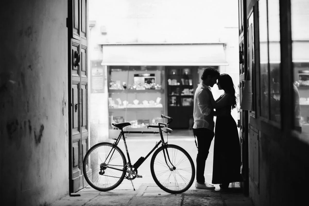 Couple in passage in front of shop window. Bicycle is in the left foreground. The picture is in black and white.