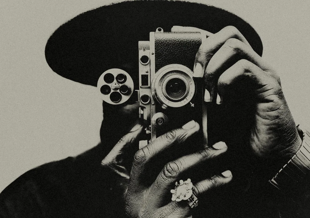 A person is holding an old camera directly in front of their face. The picture is in black and white, the camera has a round film reel element. The person is wearing a hat, a large ring and a wristwatch.