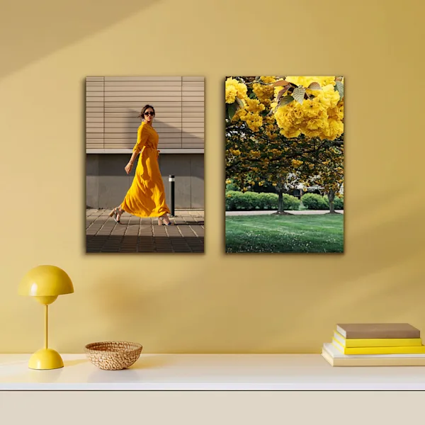 Due stampe murali verticali senza cornice visibile sono appese fianco a fianco su una calda parete gialla. Il motivo a sinistra ritrae una donna in un vivace abito giallo, quello a destra un primo piano di fiori gialli. Sulla credenza bianca sottostante, una lampada gialla minimalista, una ciotolina e una pila di libri rifiniscono armoniosamente il concetto cromatico.
