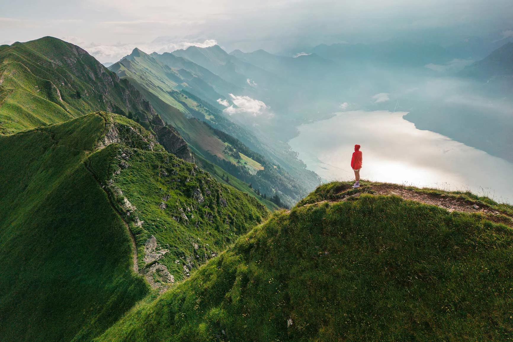 A person in a red jacket stands on a grassy mountain peak overlooking a fog-shrouded mountain range and valley.