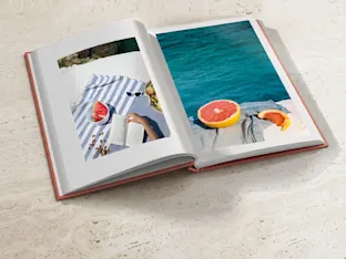 Vista superior de un libro de fotografía premium abierto con una tapa dura de color salmón, apoyado sobre una superficie de piedra de travertino claro. La página izquierda muestra una fotografía vertical de una persona leyendo un libro sobre una toalla de playa a rayas azules y blancas. La página derecha presenta un primer plano cuadrado de un pomelo cortado con el océano turchese de fondo. El diseño minimalista resalta la impresión editorial de alta gama.