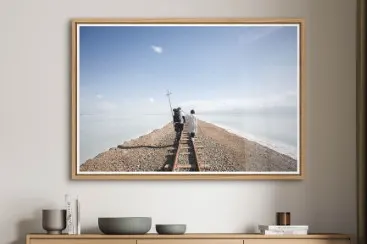 Zwei Personen gehen auf einem schmalen Damm mit Bahngleisen ins Meer unter blauem Himmel.