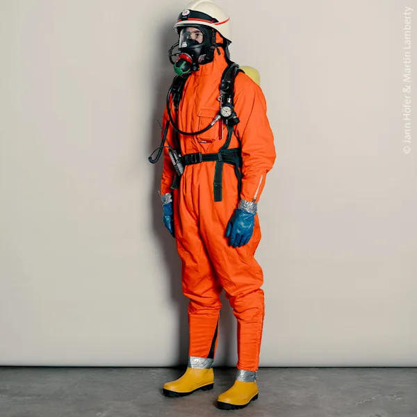 Studio portrait of a person wearing an orange protective suit, blue gloves, yellow boots, and a helmet with a gas mask. Photographed by Jann Höfer & Martin Lamberty, the image highlights safety gear typically used in industrial, emergency, or hazardous environments.