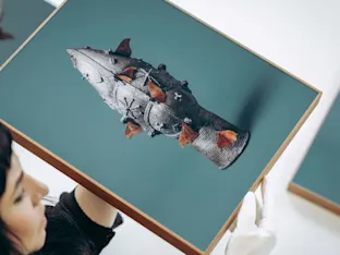 Die Nahaufnahme zeigt eine Person von der Seite im Profil, die ein querformatiges Bild in einem hellen Holzrahmen anhebt. Sie trägt weiße Handschuhe, um das Kunstwerk zu schützen, welches ein verziertes Bronzeobjekt mit braunen Faltern vor blaugrünem Hintergrund zeigt. Oben links und unten rechts im Hintergrund sind jeweils Ausschnitte weiterer, identisch gerahmter Bilder zu sehen. Die Szene dokumentiert die präzise Handhabung von Kunstfotografien in einer Galerie.