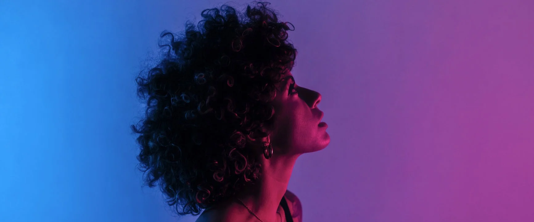 Portrait of a woman with curly hair, shown in profile against a blue-violet background with color filters.