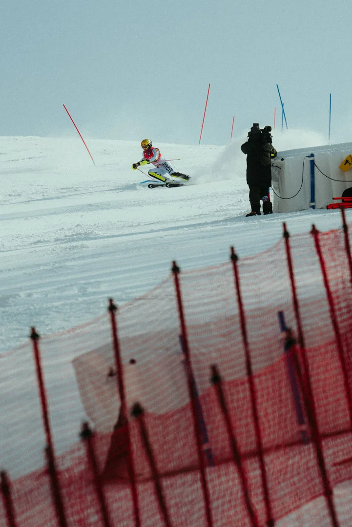 A skier in racing gear navigates down a snowy slope, passing red slalom gates. In the foreground, a red safety net is blurred, while a camera operator on the side of the track films the action.