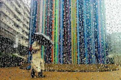 A rainy close-up with water droplets; in the background, a blurred figure holding an umbrella stands in front of a tall building with colorful vertical stripes.