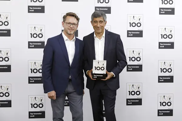 Deux hommes en costume tiennent un trophée du prix de l'innovation Top 100 devant un mur de sponsors.