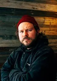 Ein Porträt von Nikita Teryoshin vor einem Hintergrund aus rustikalen Holzplanken. Er trägt eine rote Strickmütze und einen dunklen Kapuzenpullover. Er blickt mit einem ernsten, direkten Ausdruck in die Kamera. Das Licht fällt von der Seite auf sein Gesicht und betont seinen Bart.