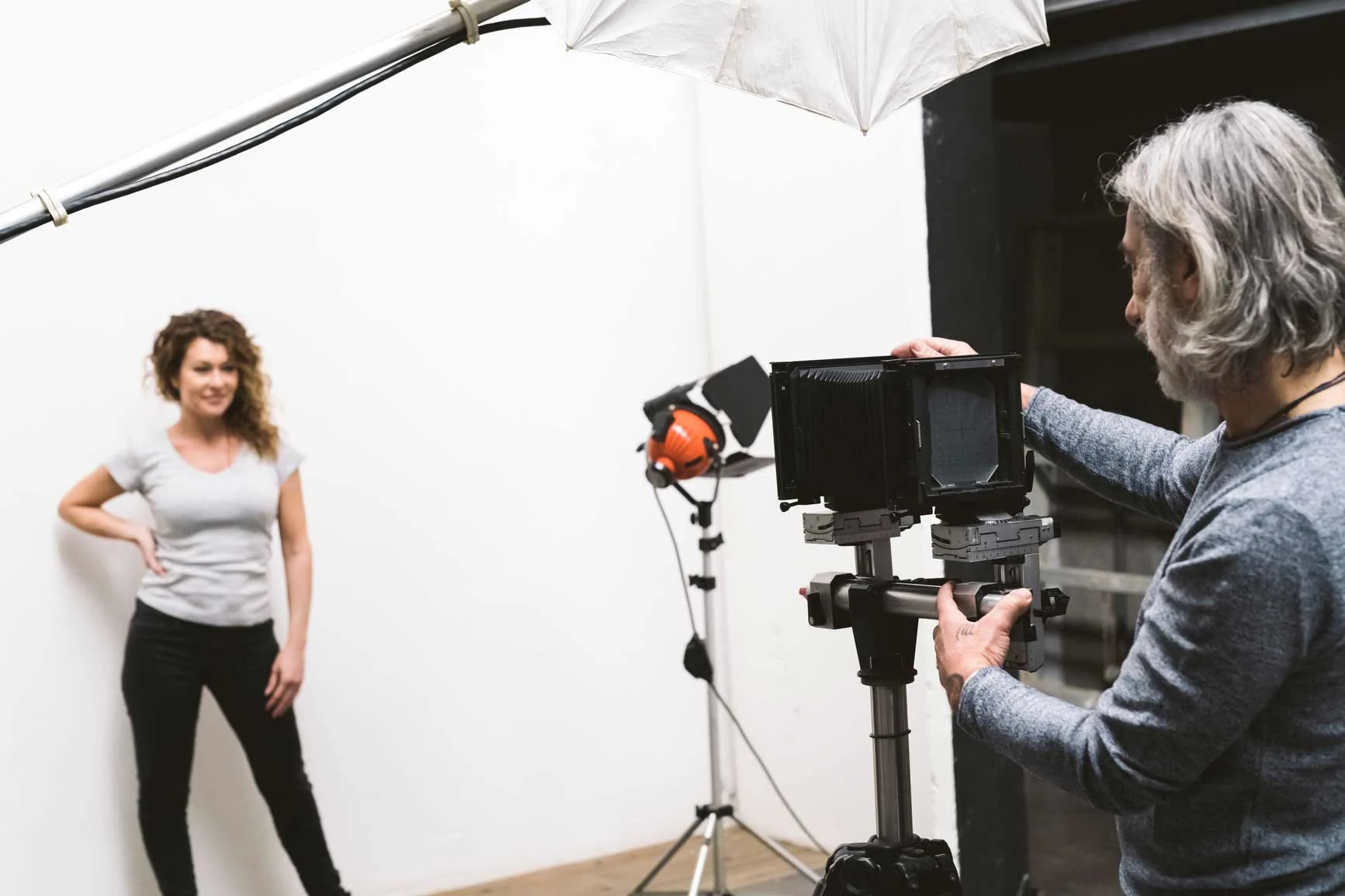 Fotograaf bedient een camera op een statief in een studio met een witte achtergrond, terwijl een vrouw poseert.