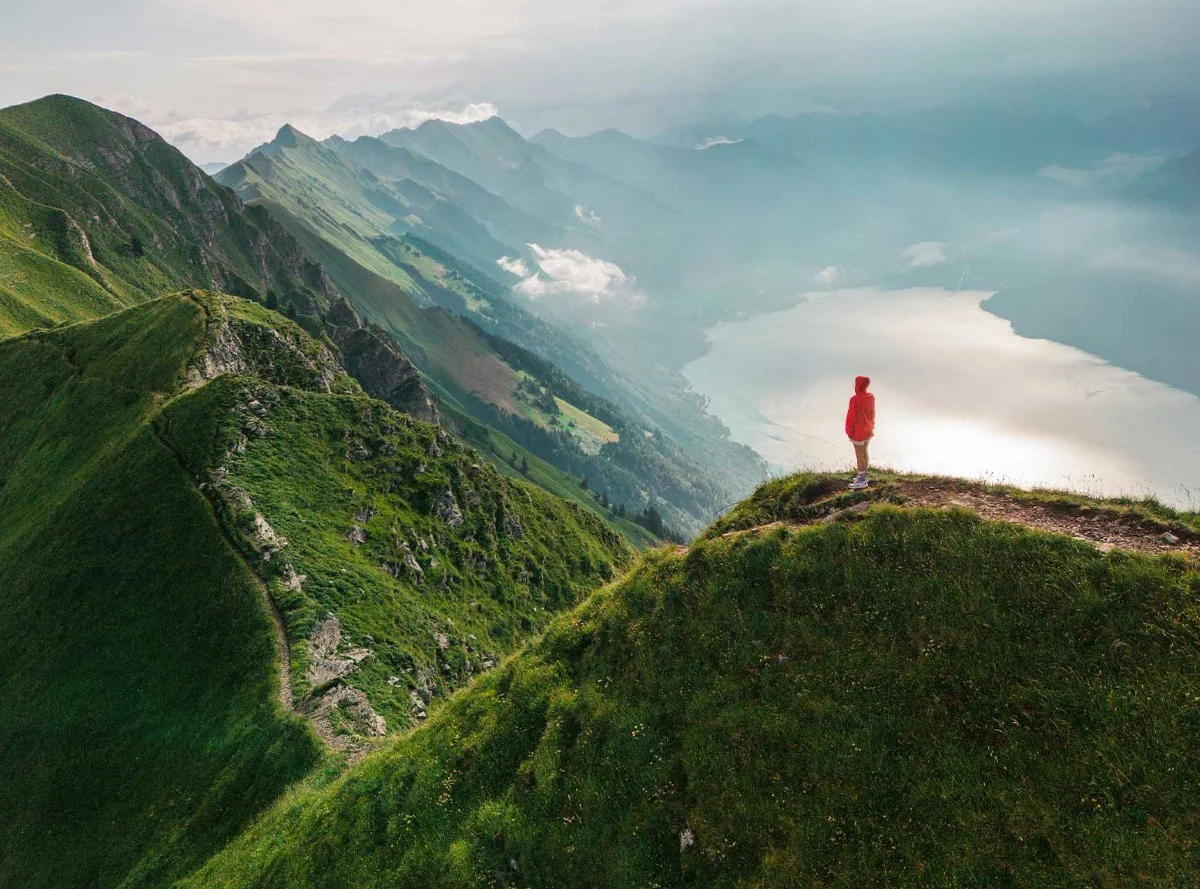 Una persona si trova sulla cima di una montagna con vista su una catena montuosa e un lago avvolti nella nebbia.