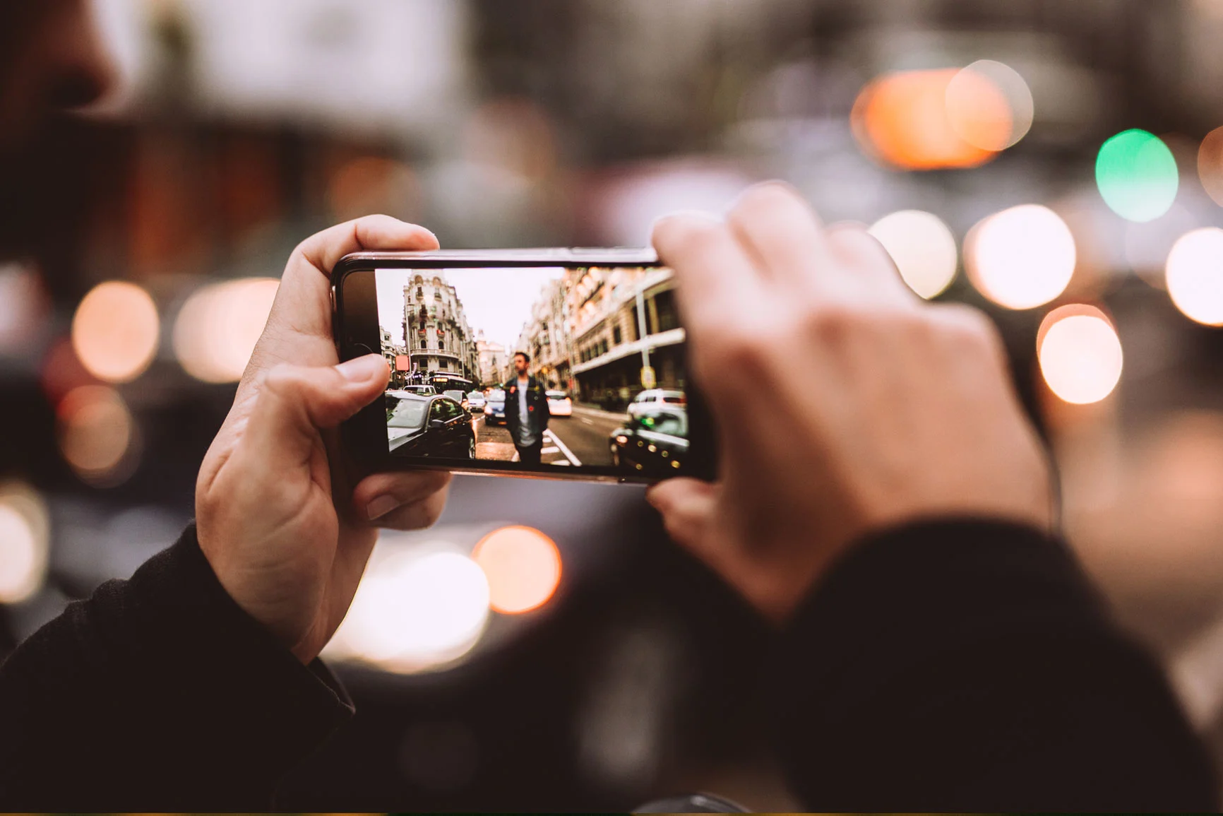 Una persona sostiene un smartphone y fotografía una calle concurrida con edificios históricos y coches.