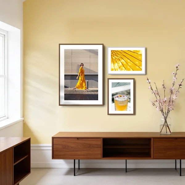 Tre immagini incorniciate di diverse dimensioni sono disposte su una parete giallo chiaro sopra una credenza in legno scuro. L'immagine più grande a sinistra ritrae una donna in abito giallo. Sulla destra, due foto più piccole sono appese l'una sull'altra: quella superiore mostra una struttura architettonica gialla astratta, quella inferiore un bicchiere con un cocktail giallo. Un vaso di vetro con rami fioriti sulla credenza esalta il tema primaverile.